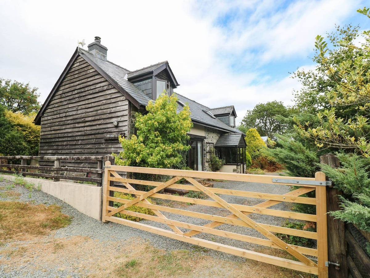 Penrhiwoli Barn timber-clad and stone cottage behind a wooden five-bar gate and gravel drive