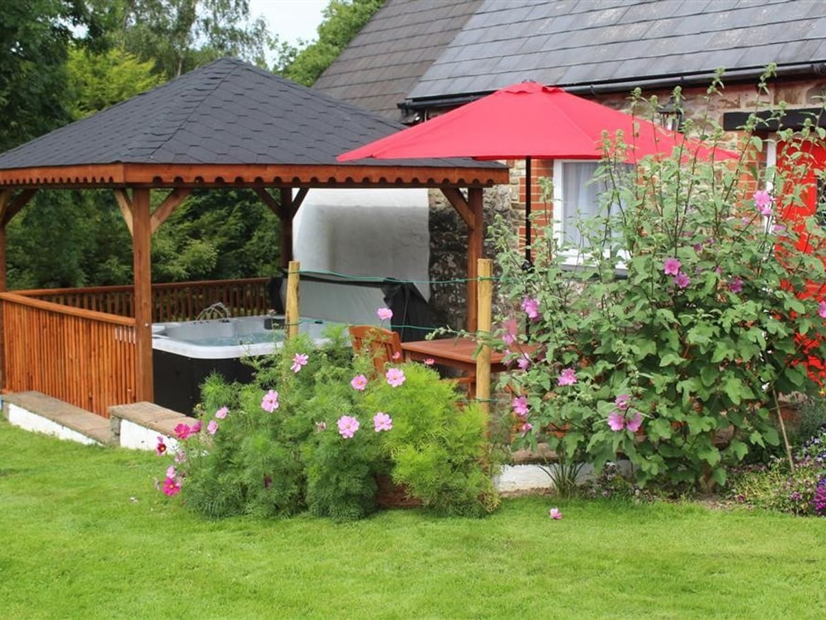 Lawn at Pond Cottage with a hot tub under a wooden gazebo, red parasol, table and flowering shrubs