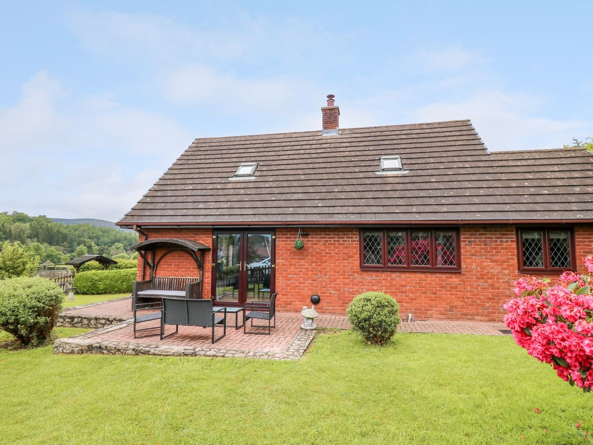 Pondarosa red brick house with a paved patio, outdoor seating and lawn, with wooded hills beyond
