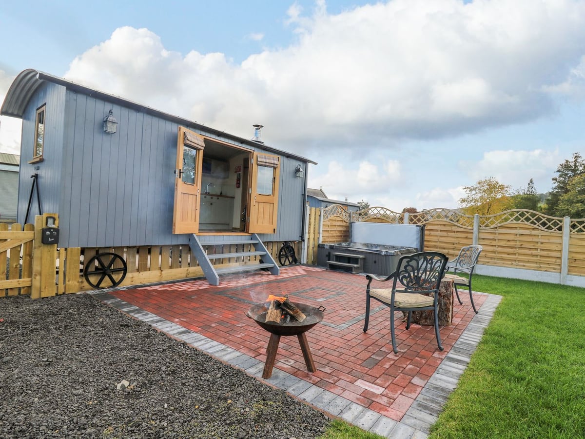 Poppy shepherd's hut with open doors, a brick patio, fire pit, chairs and hot tub beside timber fencing