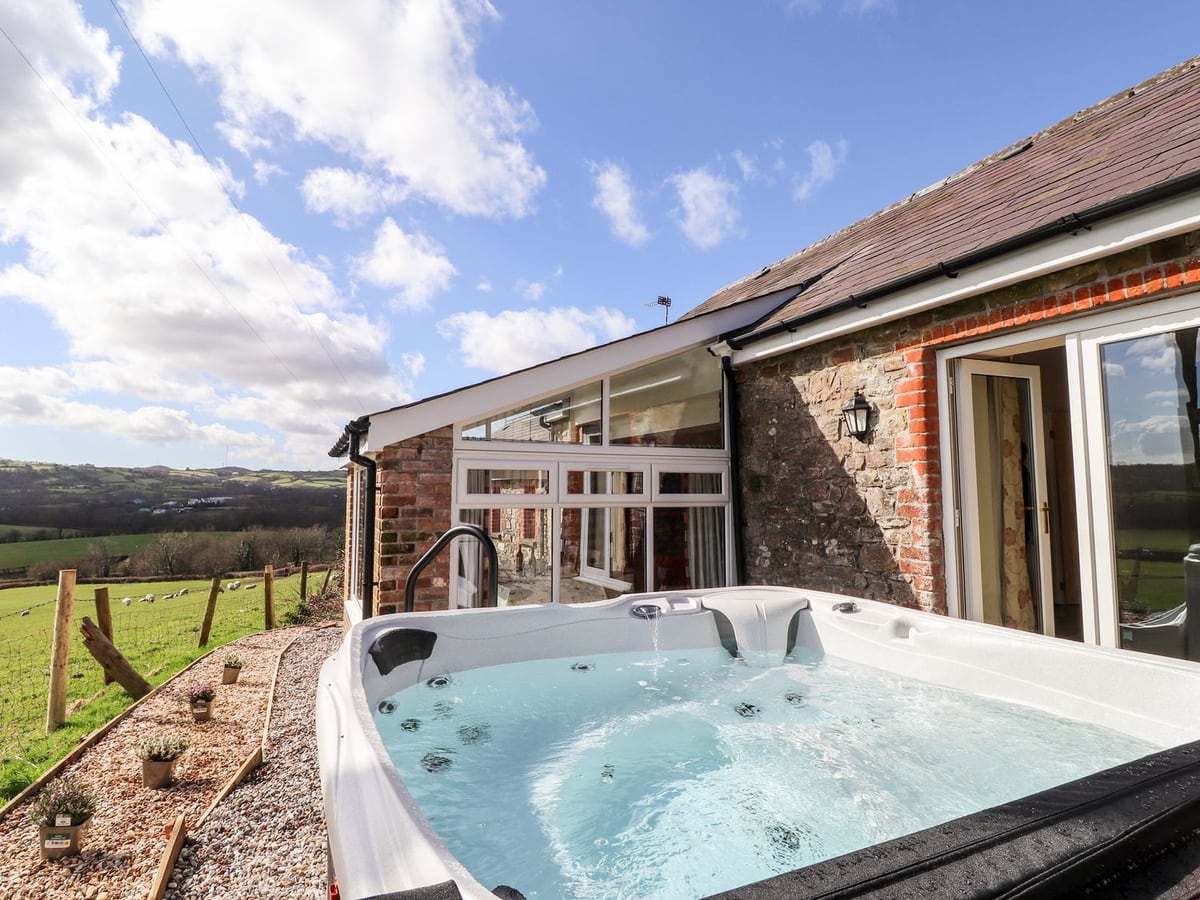 Hot tub beside Sewin Cottage's stone and brick wall and conservatory, with fields and hills beyond
