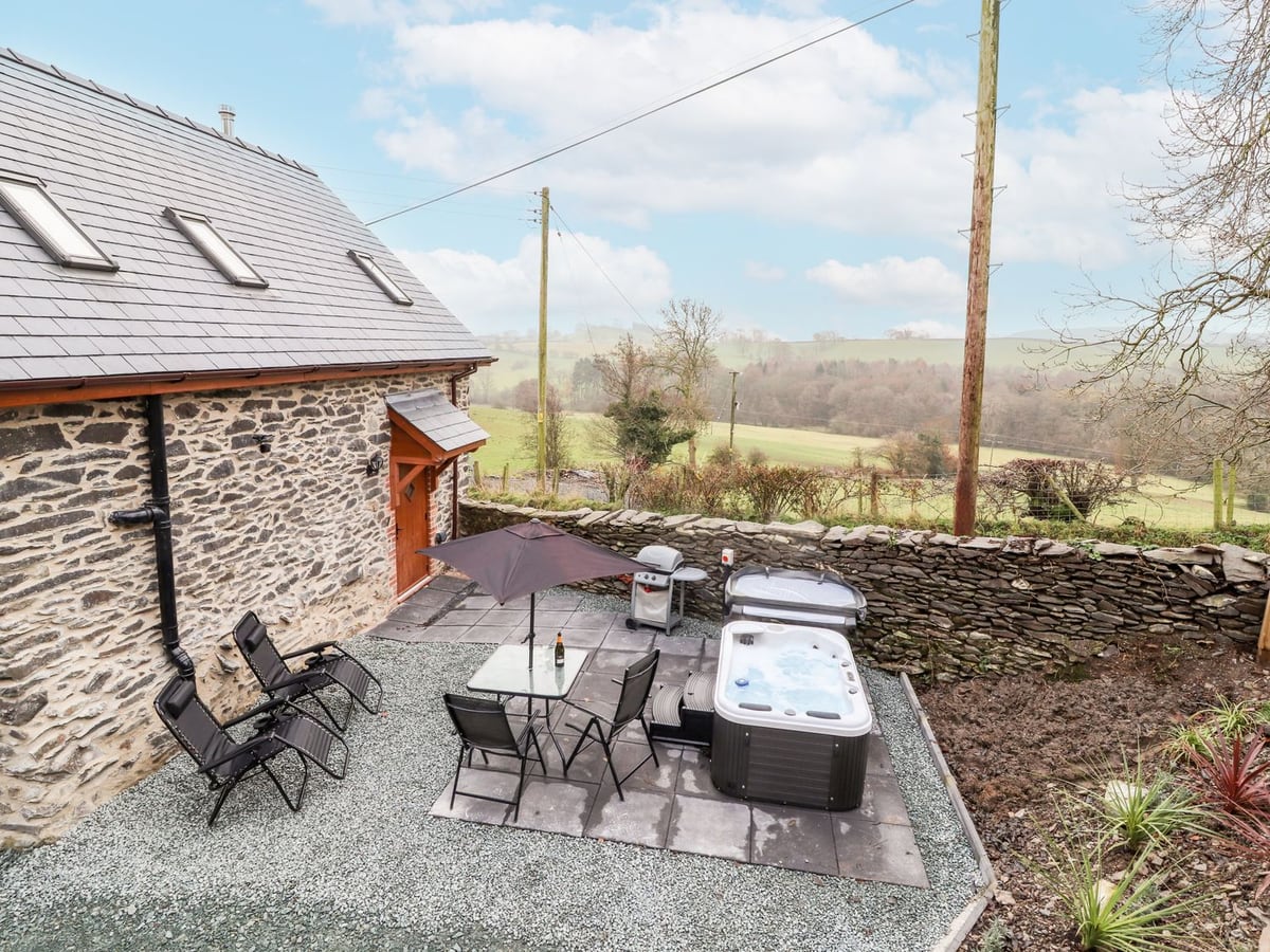 Stone barn patio at Stabl with a hot tub, table, parasol and loungers above rolling fields