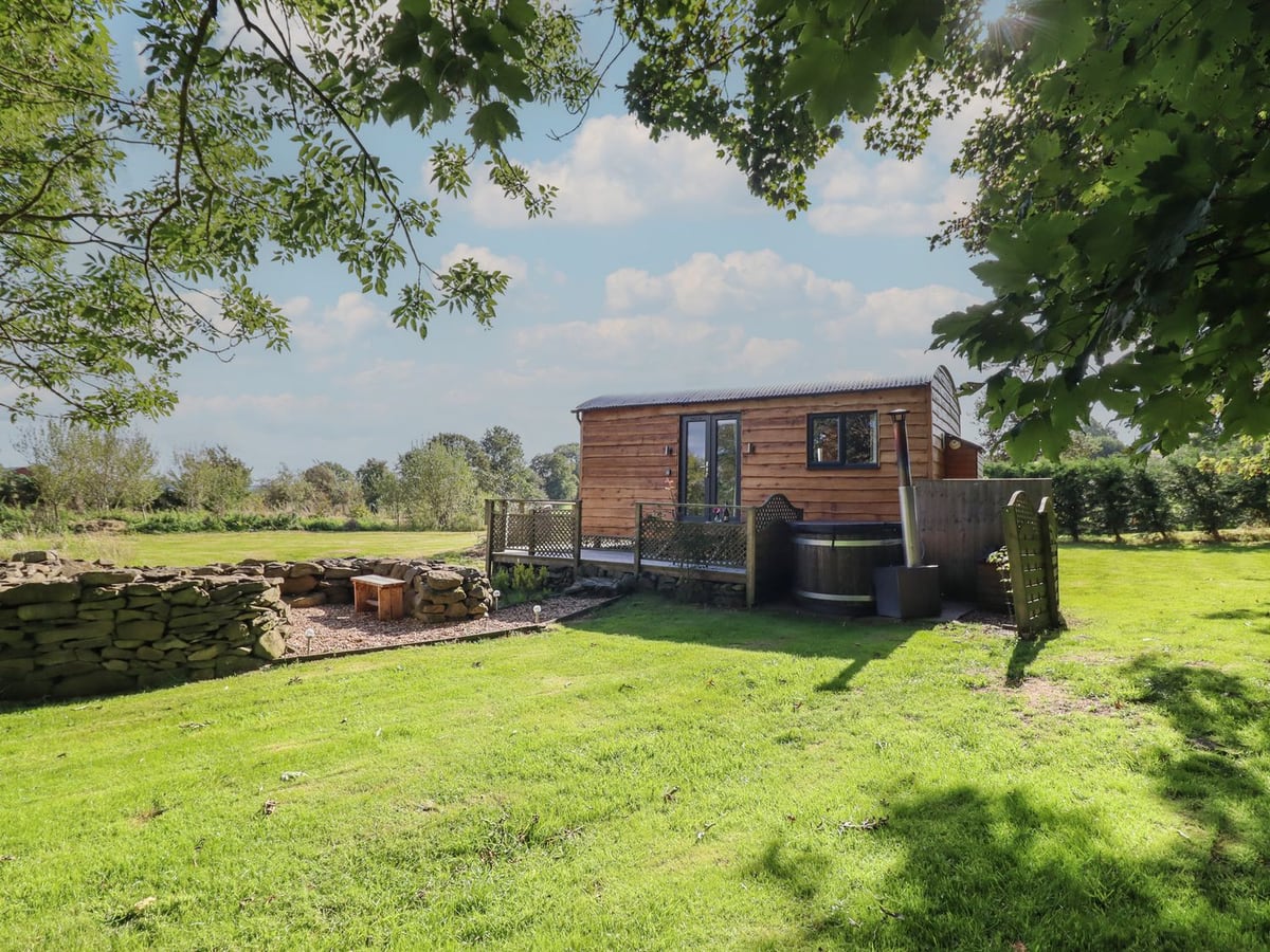 Stag Lodge Pod on a lawn with its deck, a wood-fired hot tub with its lid on and stone-walled seating