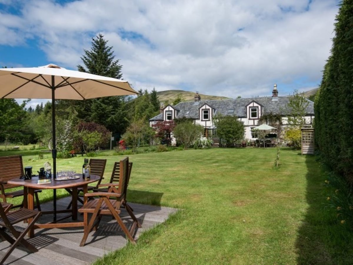 Strone House beyond a broad lawn, with a wooden dining table, parasol and mature garden trees