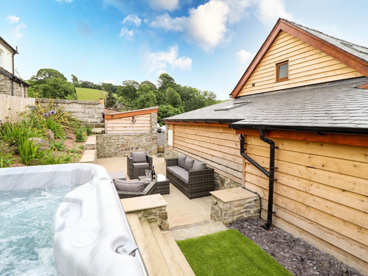 Hot tub at Swallows Rest beside a stone patio with rattan seating, timber steps and artificial turf