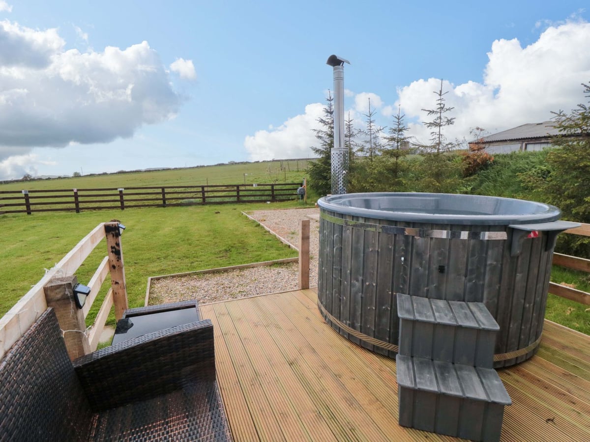 Round hot tub and wicker chairs on the deck at Swift Cabin, with grass and fields beyond