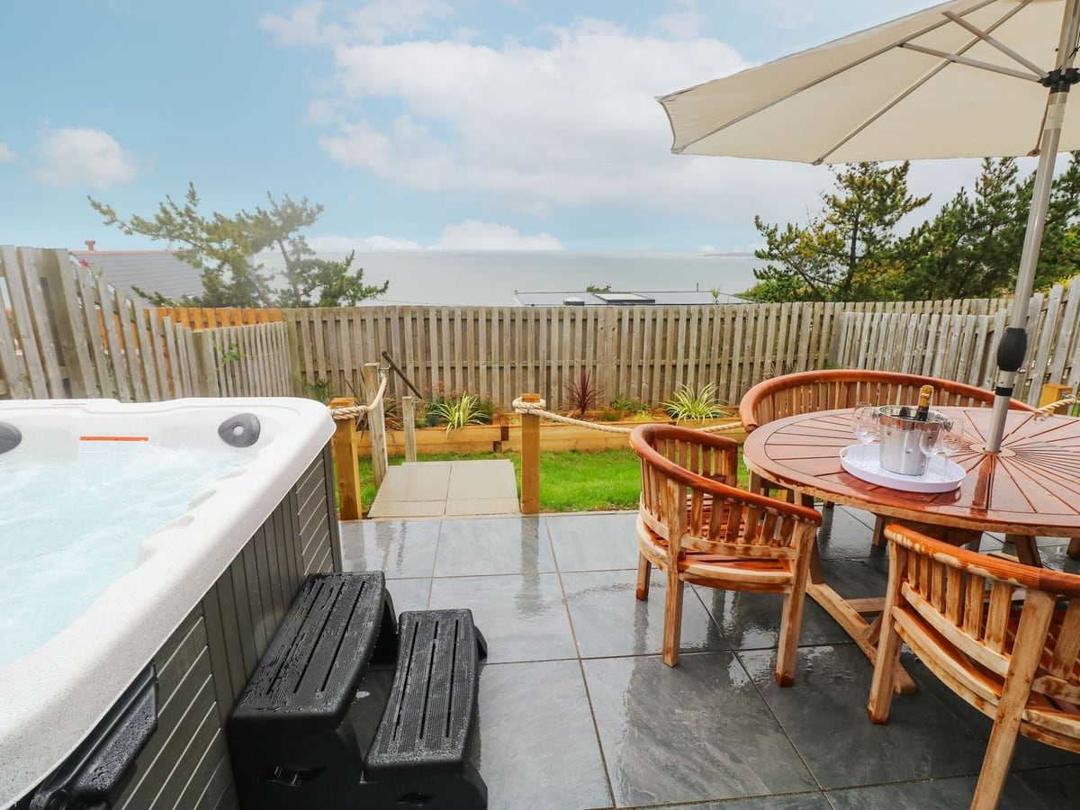 Hot tub and patio dining set at Ty Glas, with steps to a fenced lawn and sea view beyond