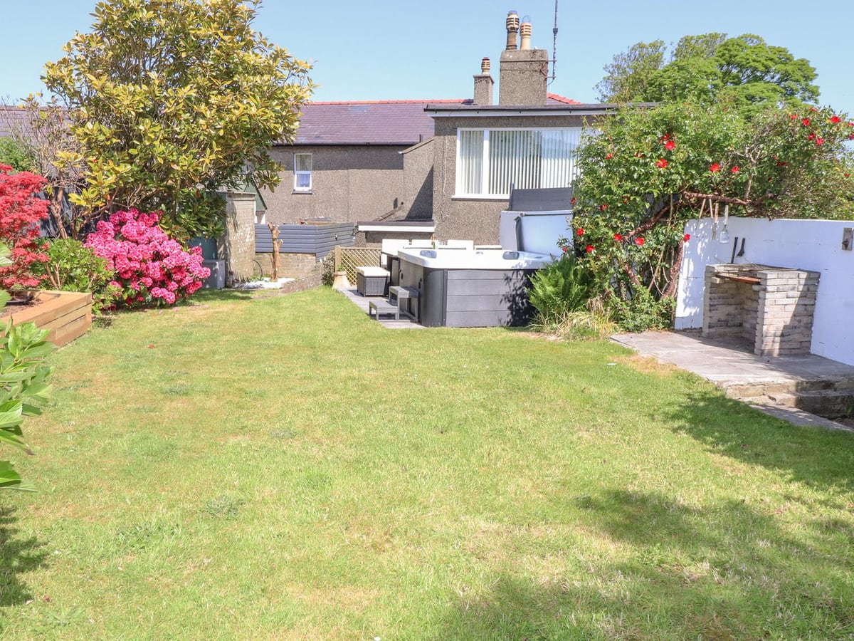 Tan Parc rear lawn with flowering shrubs, a hot tub, brick barbecue and the back of the house