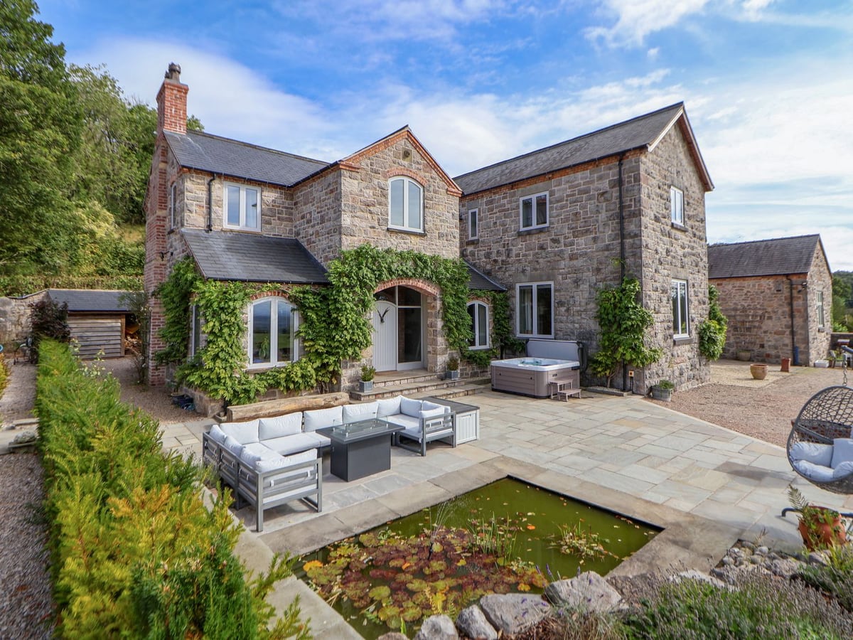 Tanat House's stone exterior behind a patio with a hot tub, seats and a small pond