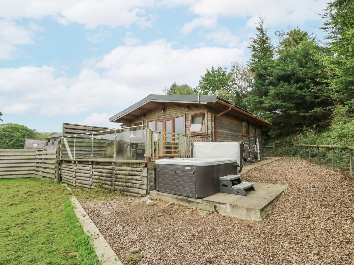 The Cabin log cabin with a glass-railed deck, open hot tub on a paved pad and grass alongside