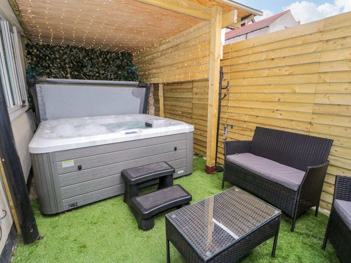 The Causeway hot tub under a timber canopy in a fenced courtyard with artificial turf and rattan seating