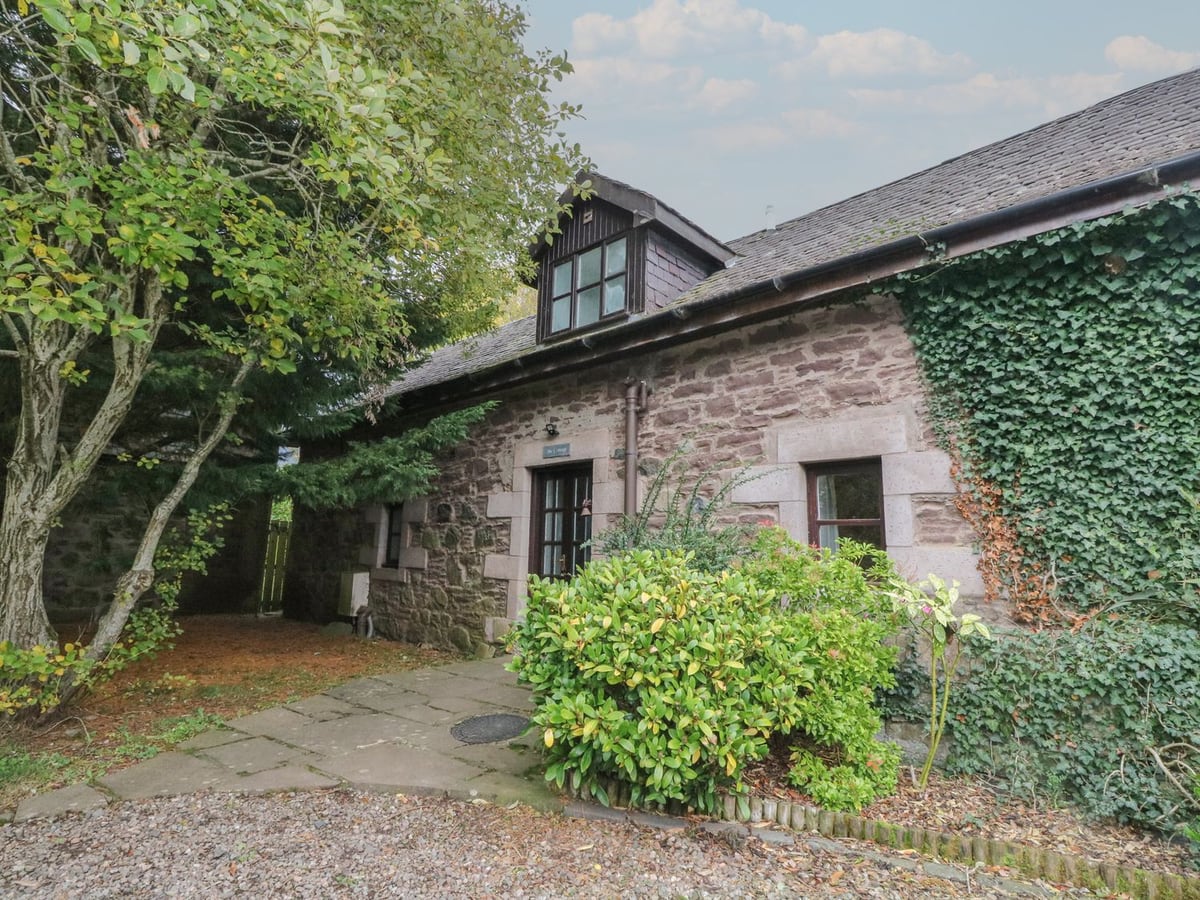 Stone front of The Cottage, with ivy, shrubs and a paved entrance beside the gravel approach
