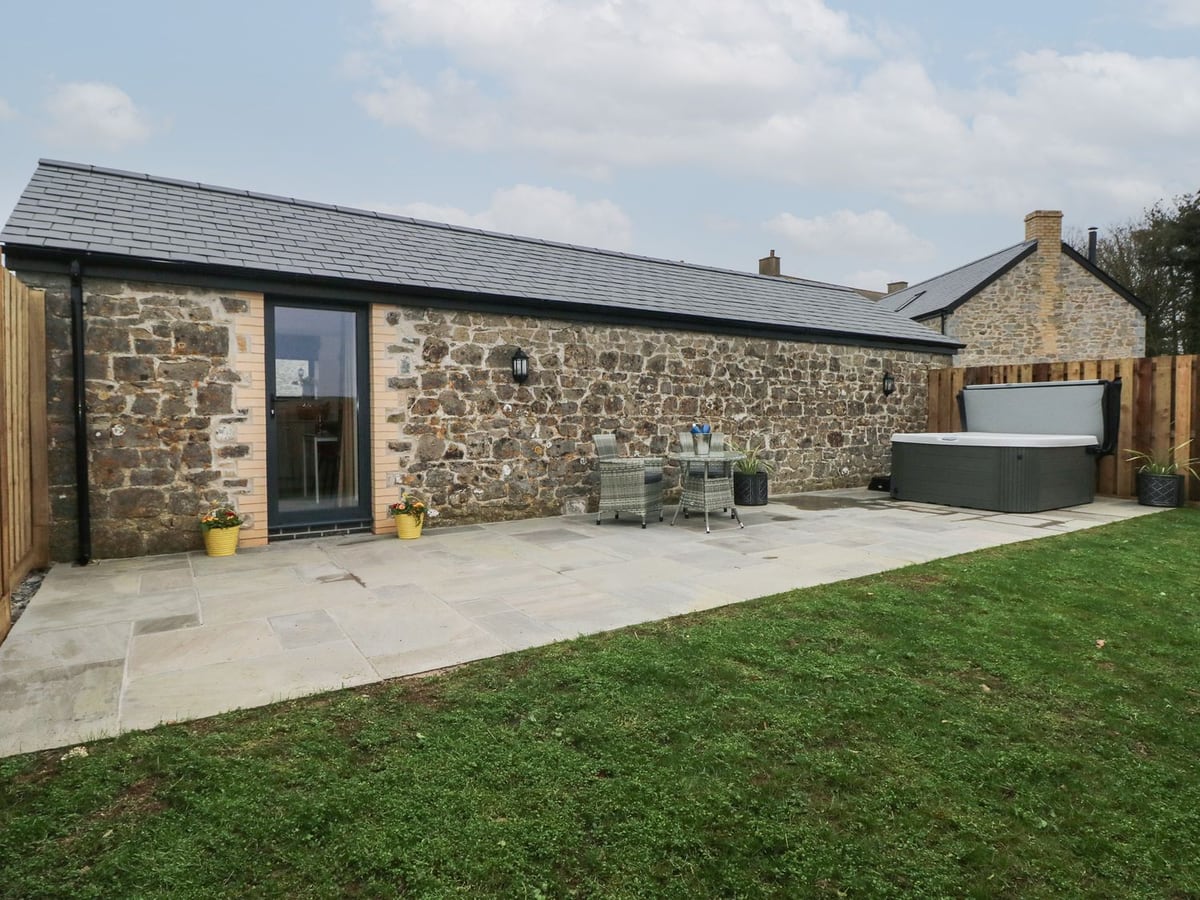 Stone barn at The Cwtch with a lawn, paved patio, bistro table and hot tub beside a wooden fence