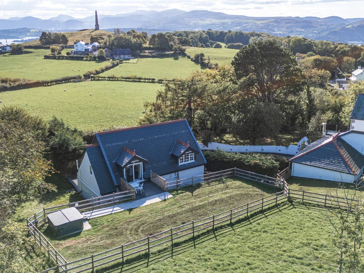 Aerial view of Gatekeepers Cottage with rail-fenced lawn, deck, hot tub with its lid on and fields beyond