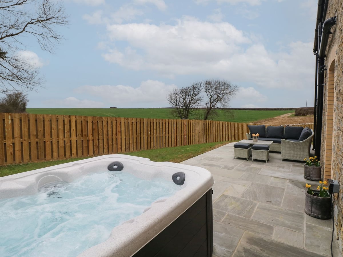 Hot tub on a paved patio at The Granary, with outdoor sofa, lawn, wooden fence and fields beyond