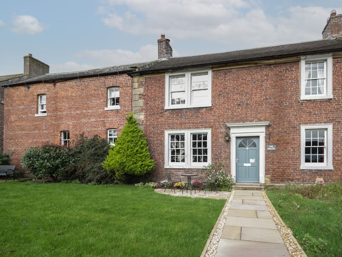 The Hollies brick frontage with a lawn, paved path and table beside the blue-grey front door