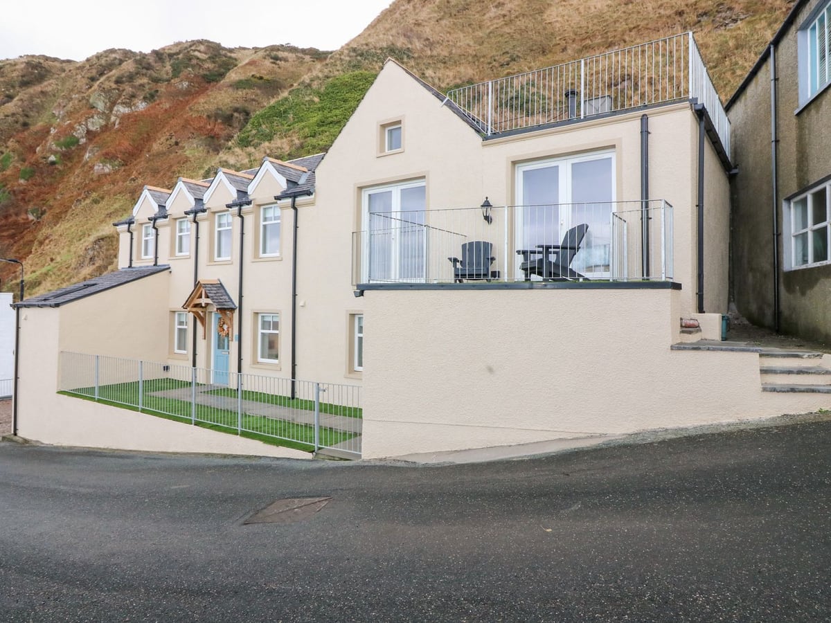 The Old Kipper Hoose beside the road, with pale walls, balconies and a hillside behind