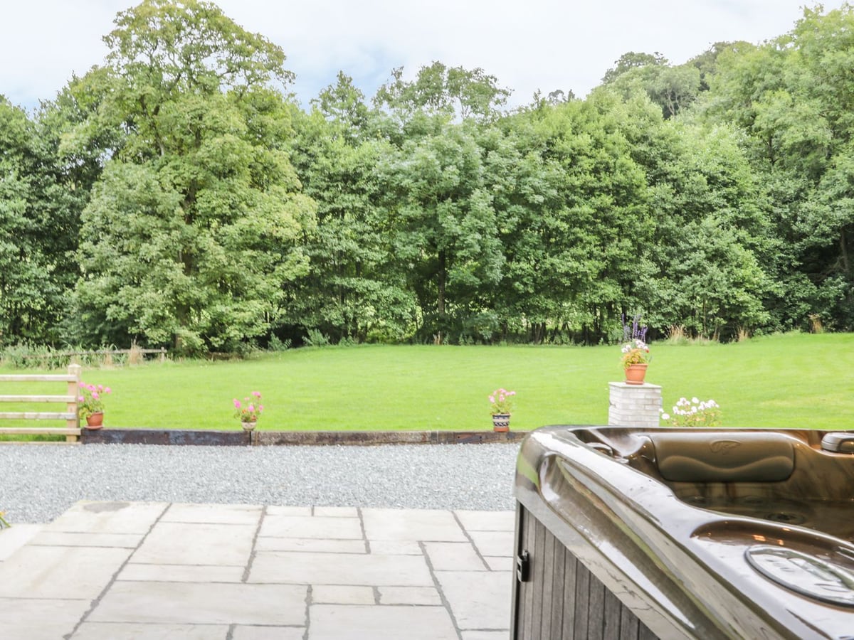 The Old Mill paved terrace with part of a hot tub, a gravel strip and a wide lawn backed by trees