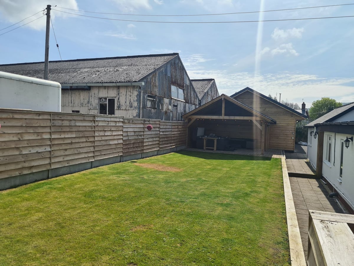 Fenced lawn at The Old Post Office leading to a timber gazebo, with large sheds beyond the fence