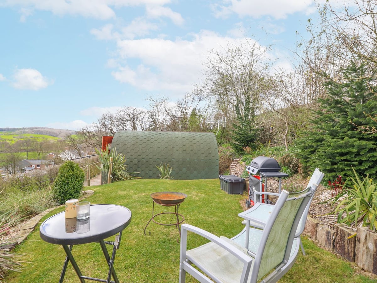 The Rhiw lawn with a fire pit, garden chairs and barbecue, the green pod behind and valley views beyond