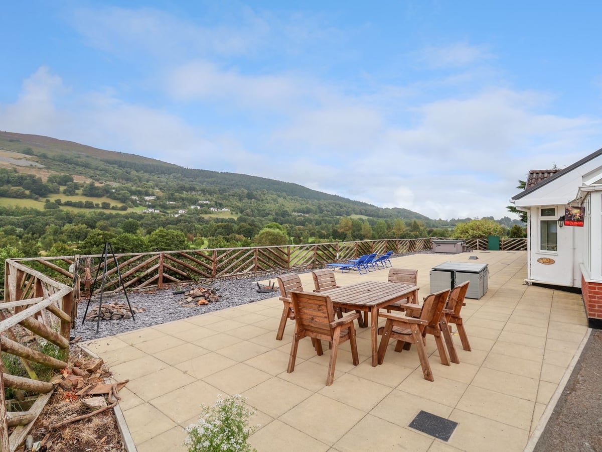 The Willows paved terrace with wooden table and chairs, loungers, hot tub and wooded valley view