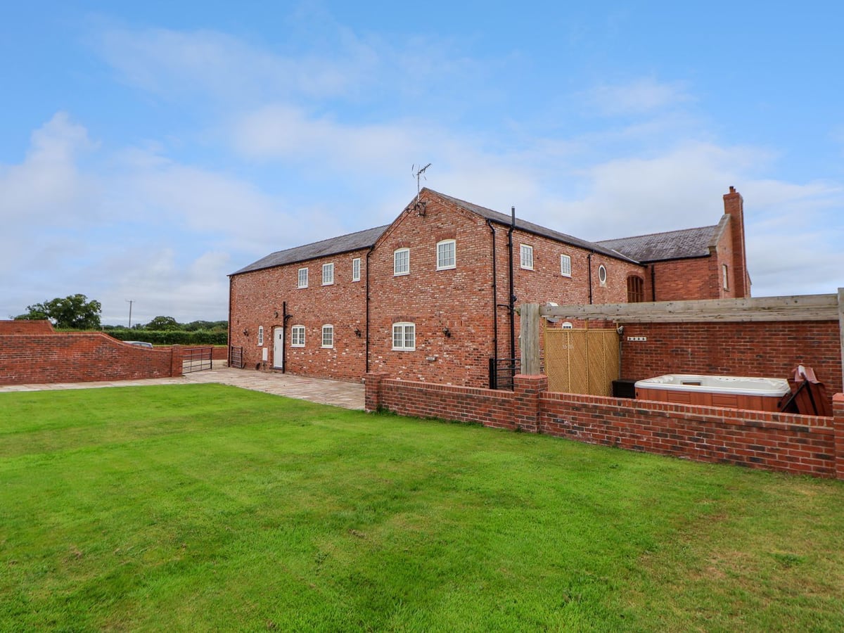Red brick barn at The Wing with a lawn and an open hot tub behind a low brick wall