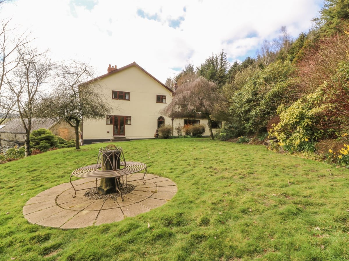 Treboeth farmhouse seen across a sloping lawn with a round tree bench, shrubs and a wooded bank