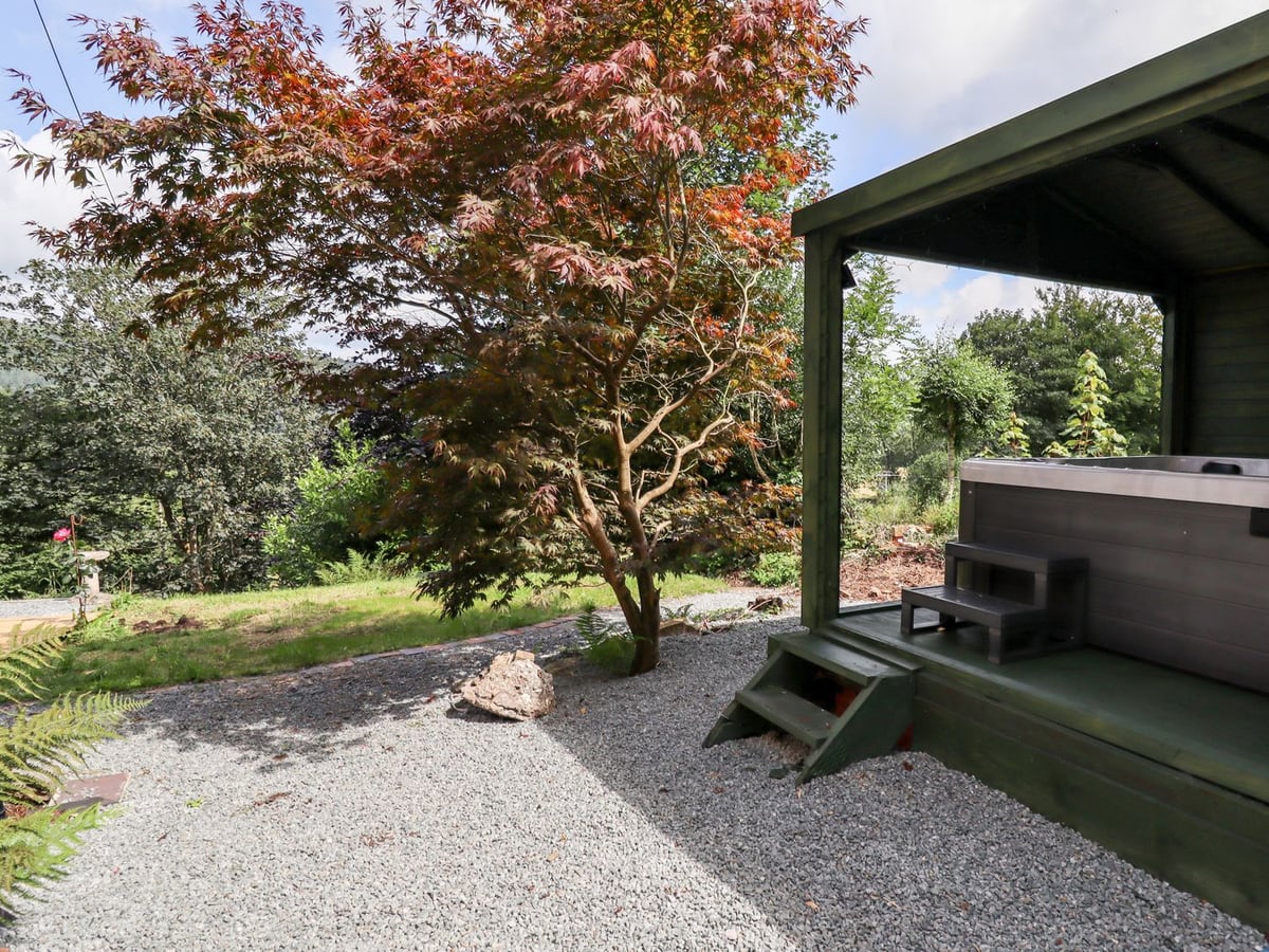 Hot tub under a green timber shelter beside a gravel path, lawn and red-leaved tree at Trem Y Don