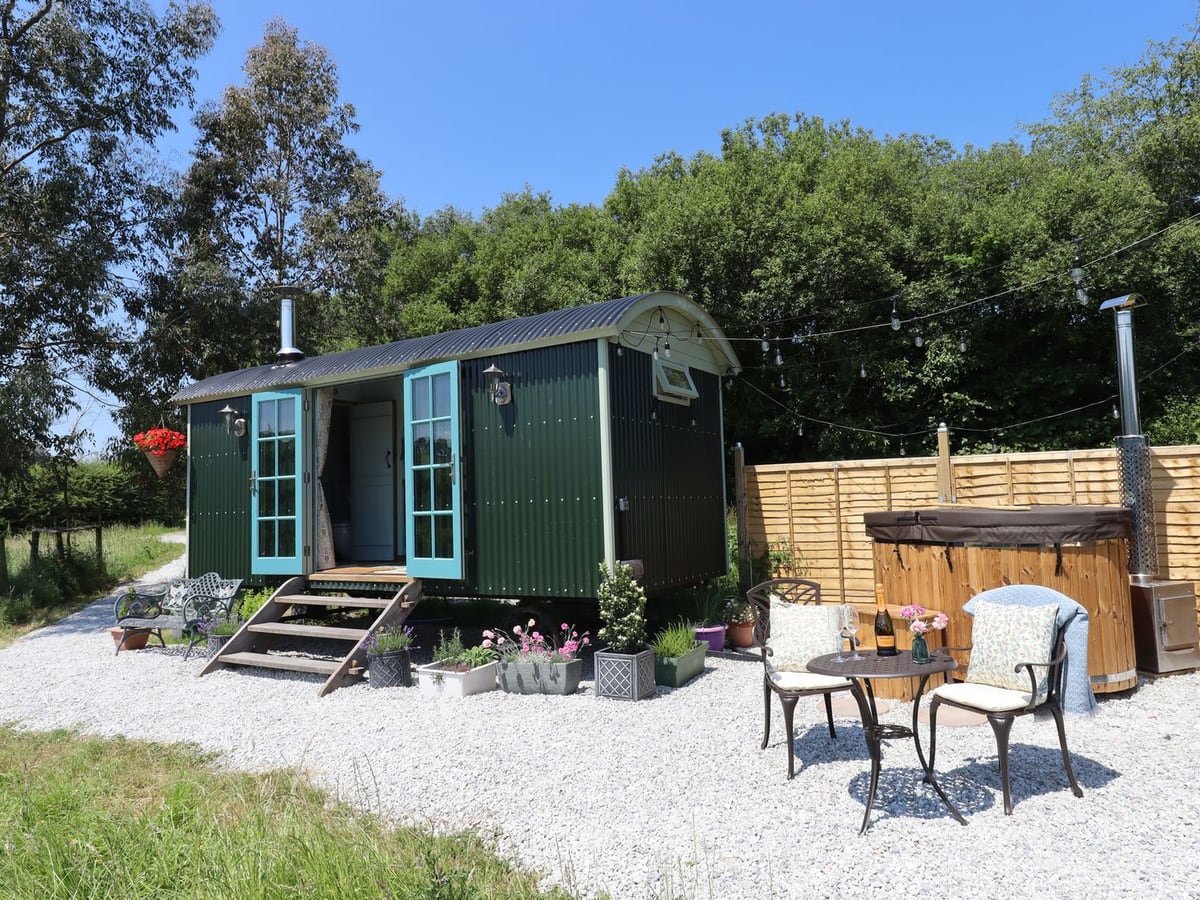 Green shepherd hut with entrance steps and a round timber hot tub at Two Moors Shepherds Hut