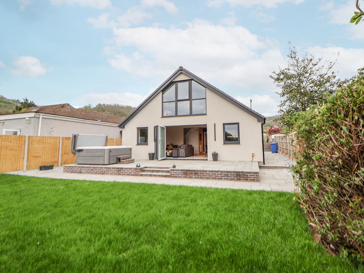 Rear of Ty Blodau with a lawn, raised paved patio, hot tub and open bifold doors to the living room