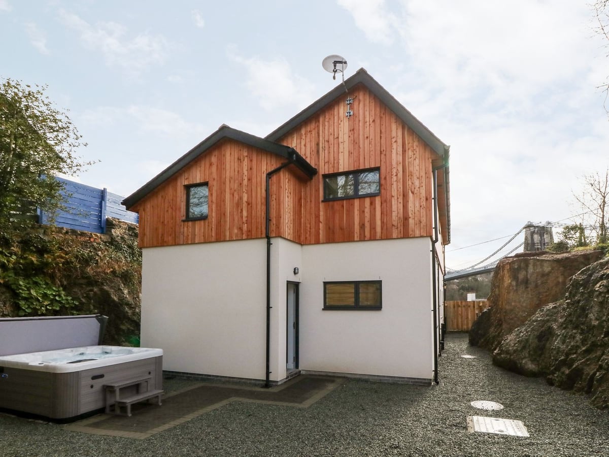Ty Chwarel with an open hot tub on gravel beside the house, a rock face and suspension bridge beyond