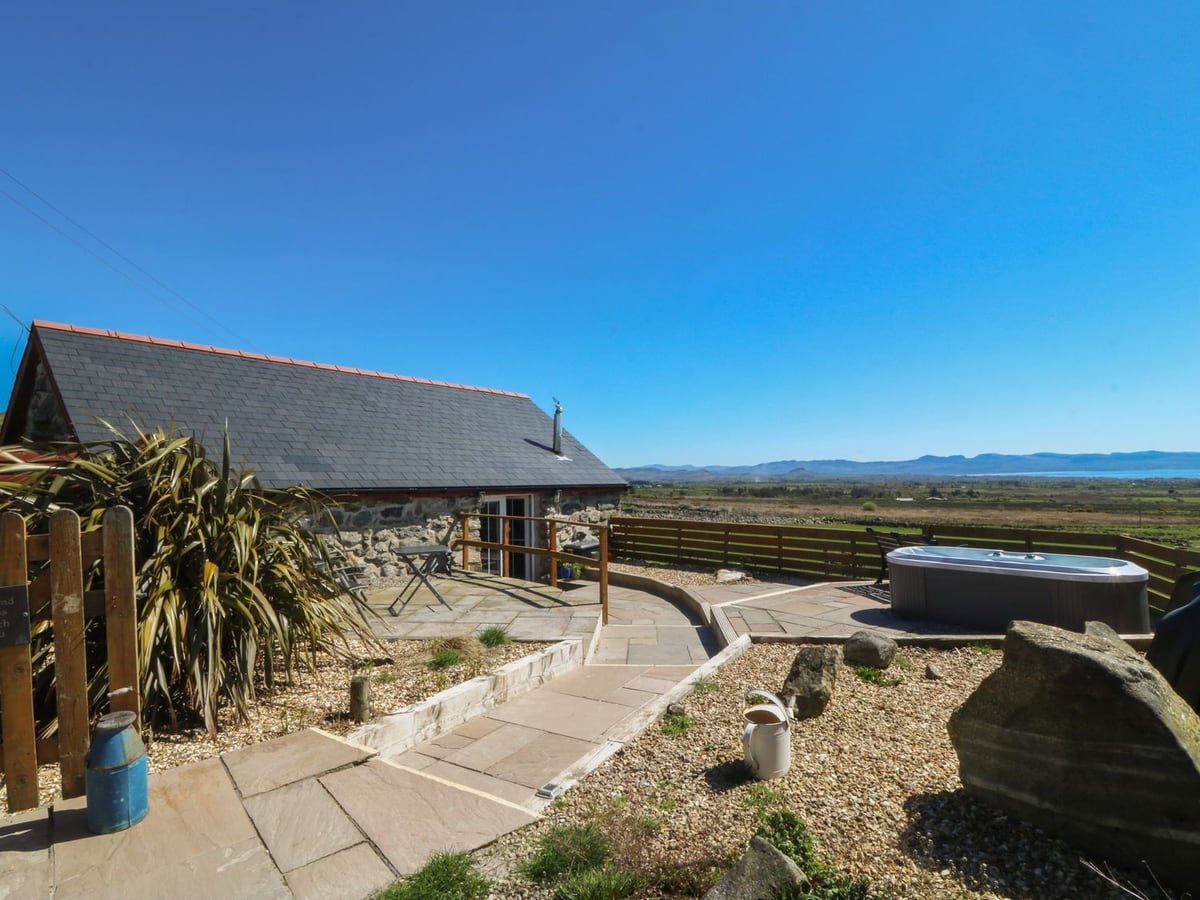 Ty Coch stone cottage with a paved patio, gravel beds and hot tub with its lid on, facing hills and sea