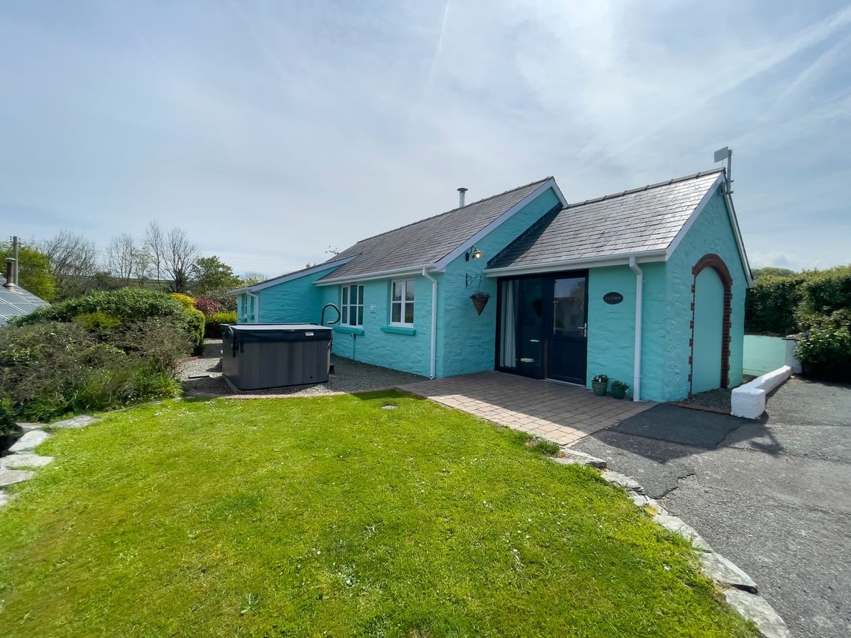 Ty Coed turquoise bungalow with a lawn, paved area by the door and hot tub with its lid on beside shrubs