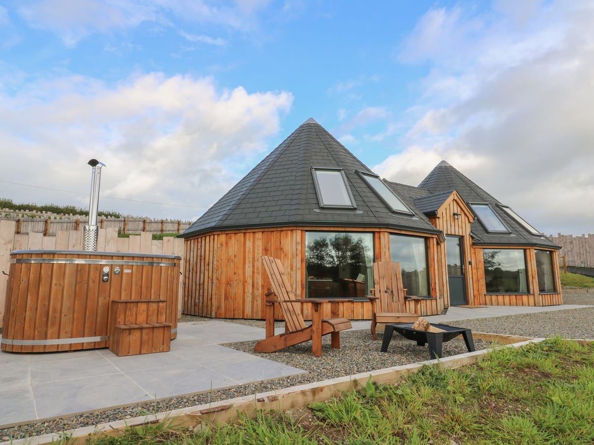 Ty Crwn roundhouse with a wood-fired hot tub on a patio, two wooden chairs, fire pit and fenced grounds
