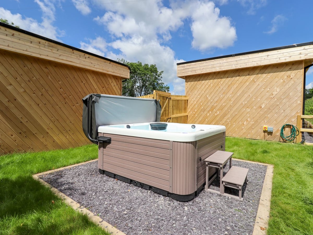 Hot tub with its lid raised on a slate gravel pad in a lawned garden between two timber lodges