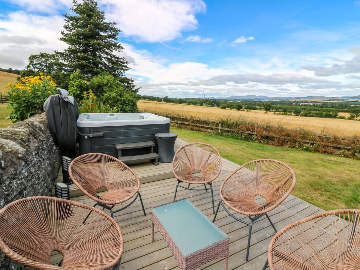 Hot tub and outdoor chairs beside a stone wall, lawn and fields at Whitehouse Woodwrae