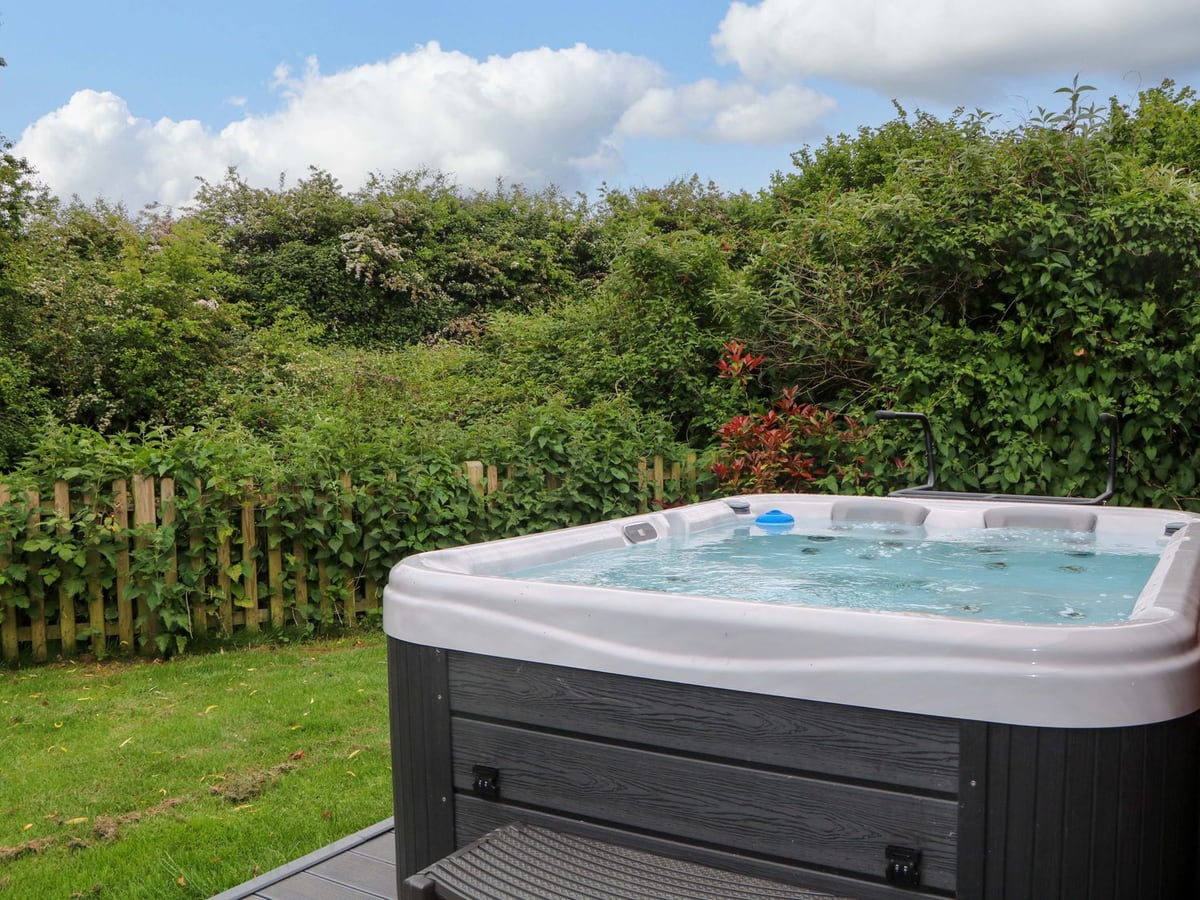 Willow Pod hot tub beside a patch of lawn, with a low picket fence covered by leafy shrubs