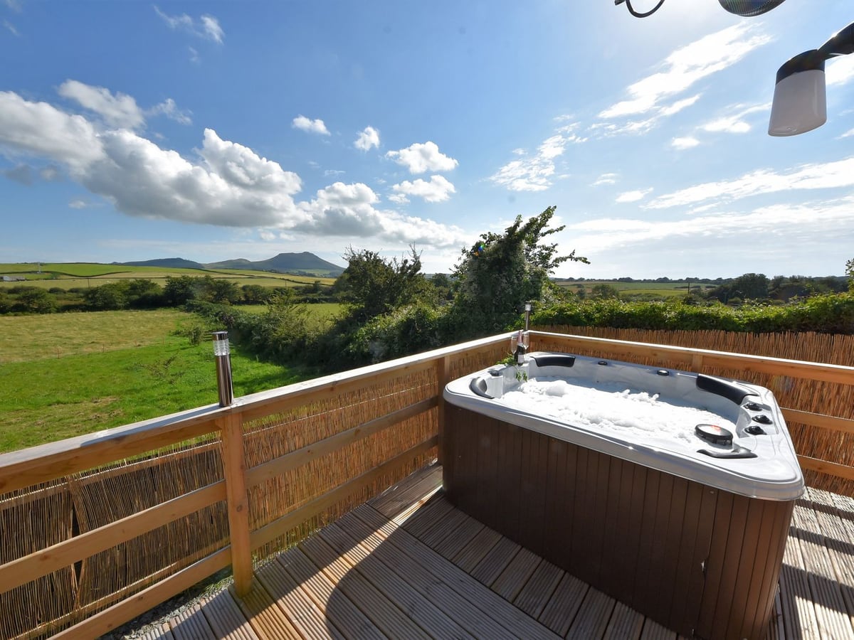 Y Caban Clyd hot tub on a timber deck with reed-screened rails, looking over fields to distant hills