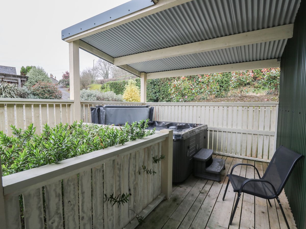 Y Felin Moana hot tub with its lid raised on a roofed deck with timber railings, a chair and shrubs