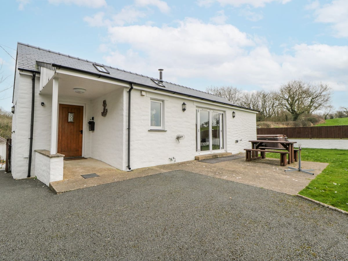 Y Felin's white rendered side with porch, patio picnic bench, hot tub with its lid on, lawn and gravel drive