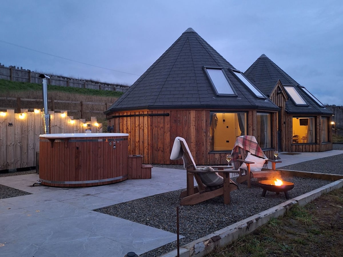Yr Awen roundhouse at dusk with wood-fired hot tub, paved patio, fire pit and two wooden chairs