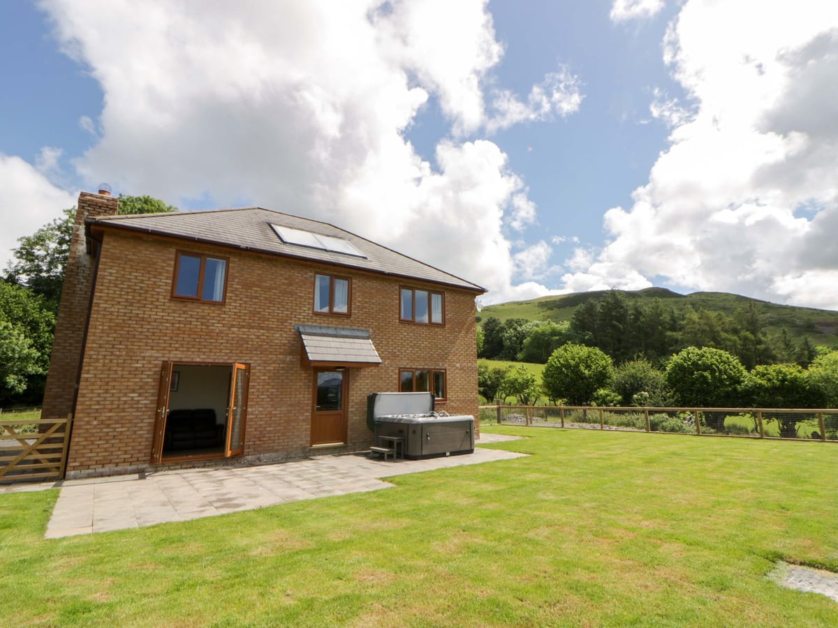 Yr Onnen brick house with a lawn, patio, hot tub with its lid raised and a fence below a green hill