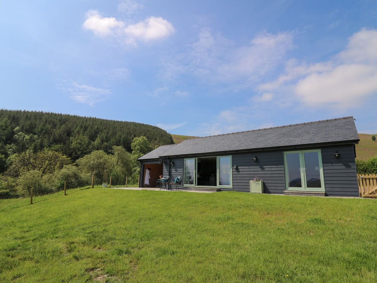 Ystrad Wen single-storey dark clad cabin with a sloping lawn, wire fencing and wooded hills behind